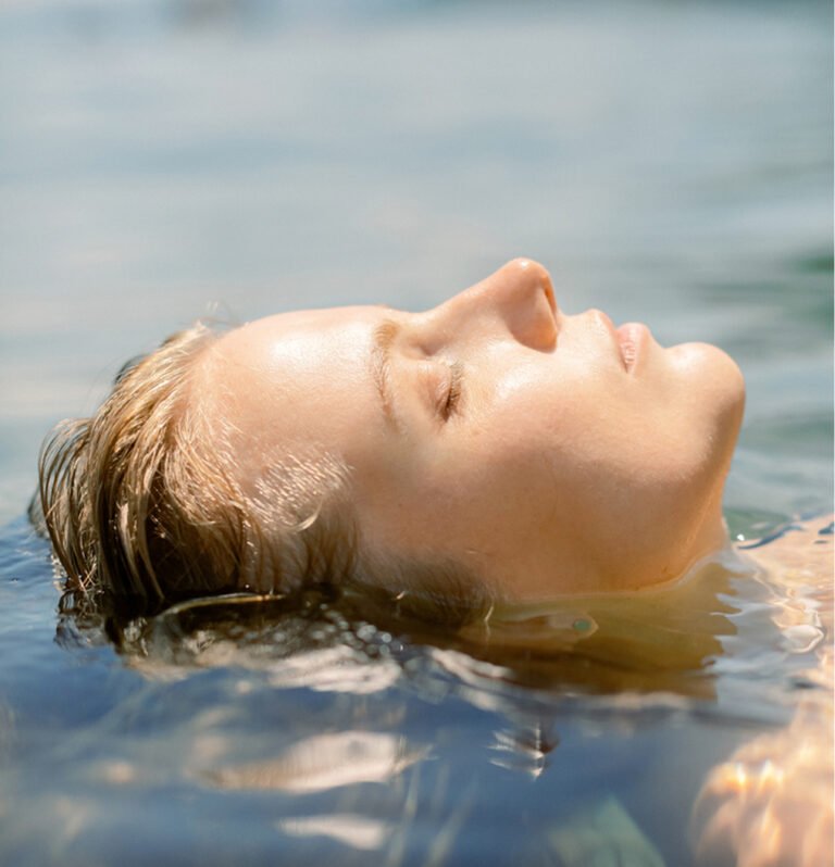 Close-up of a woman floating peacefully on her back in calm blue water with her eyes closed.